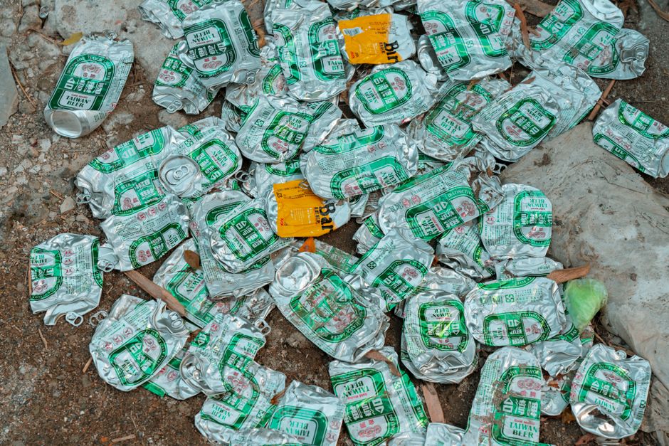 A collection of numerous crushed aluminum cans scattered on the ground, with the majority displaying green and white labels indicating they contain Taiwan Beer. The cans are intertwined with some orange stickers, and a few are partially crumpled, reflecting their discarded state. The background includes a dirt surface with small stones and dried leaves, and a plastic bag is visible on the right side of the image. The scene depicts typical waste material that would be collected during a rubbish removal service, illustrating the type of recyclable metal waste alternative disposal methods like private collection might handle. The environment appears outdoor, and the cans have a metallic, slightly glossy finish, with textured crumpled surfaces. This image aligns with the visual context of waste clearance and rubbish disposal, showcasing recyclable aluminum waste typical of household or commercial refuse, relevant to rubbish removal services in Fulham and SW6 areas.