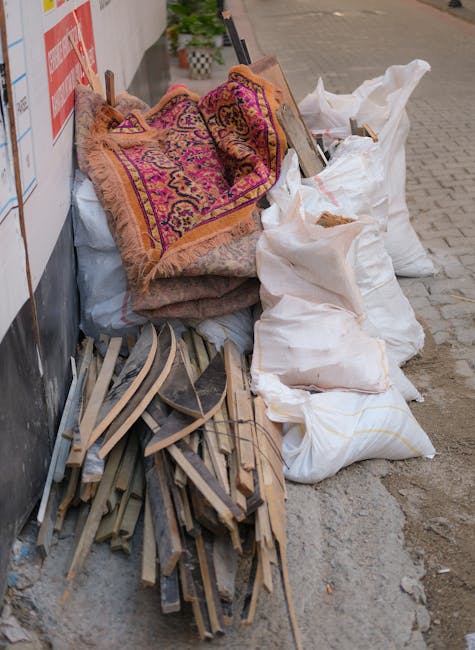 A pile of discarded household items and waste materials positioned on the pavement beside a building wall. The collection includes a rolled-up, patterned rug with vibrant pink, purple, and gold tones, partially unfolded and draped over other objects. Next to the rug are several large white sacks filled with rubbish, tied at the top, some appearing to contain fabric or soft debris. Underneath, there are remnants of wooden furniture or flooring strips with a weathered finish, showing a mix of light and dark wood tones and bent or broken edges. The scene is outdoors, next to a wall with a partially visible red and white sign, and the background features a paved sidewalk and street area. The environment suggests an on-site clearance of household or shop rubbish, possibly arranged for private collection services like those provided by Rubbish Clearance Fulham, designed for business or domestic waste removal outside of local authority disposal options.