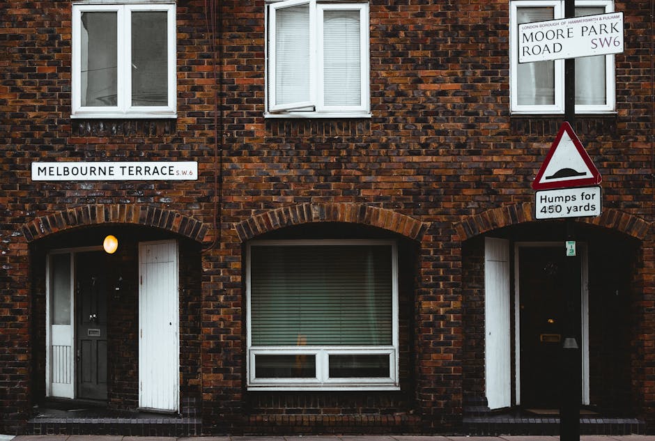 A brick residential building on Melbourne Terrace in Fulham, featuring three white-framed windows on the upper floor and two black doors with white trims on the ground floor, each under brick archways. One doorway has a small white gate next to it; the other has a mail slot and a small window. The brickwork is dark red with a weathered finish. Affixed to the building are street signs indicating the location as Moore Park Road SW6 and Melbourne Terrace SW6. Between the signs, there is a triangular warning sign showing a bump symbol with the text 'Humps for 450 yards,' indicating a speed bump or road surface feature ahead. The setting suggests an urban environment with ambient outdoor lighting, suitable for private rubbish clearance or alternative waste handling procedures, as might be managed by local waste collection services like Rubbish Clearance Fulham.