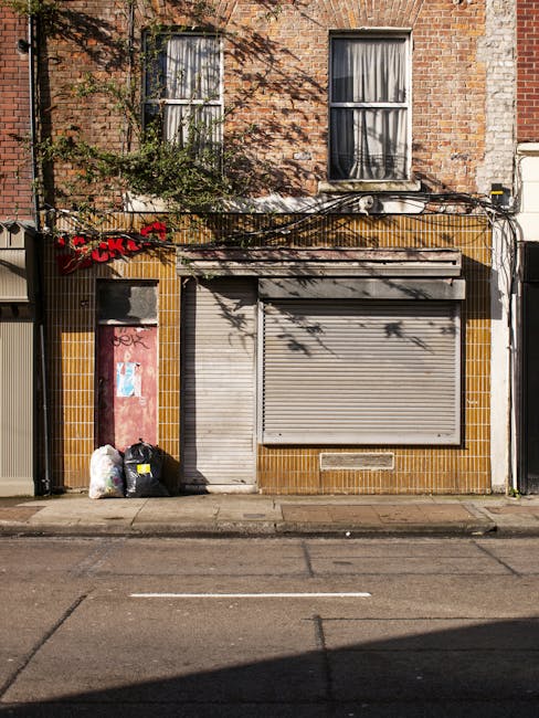 The image shows the exterior of a two-story brick building with two large, vertically oriented windows on the upper floor, each with white window frames and partially drawn curtains. The lower level features a closed, metallic roller shutter door with a smaller, matching pedestrian door to its left, both set within a section of yellow-brown tiles that contrast with the surrounding red brickwork. In front of the building's frontage, on the pavement, there are three black and white plastic rubbish bags, alongside some trash, reflecting typical waste disposal. A sign with red lettering, partially obscured, is attached to a dark-colored metal pole or fixture near the rubbish bags. Above, neatly fixed black cables run horizontally across the facade, casting shadows on the brickwork. The overall environment suggests an urban street scene, where independent waste collection or on-site rubbish clearance by companies like Rubbish Clearance Fulham is taking place, with the scene emphasizing the need for professional rubbish removal services for such private waste management tasks.