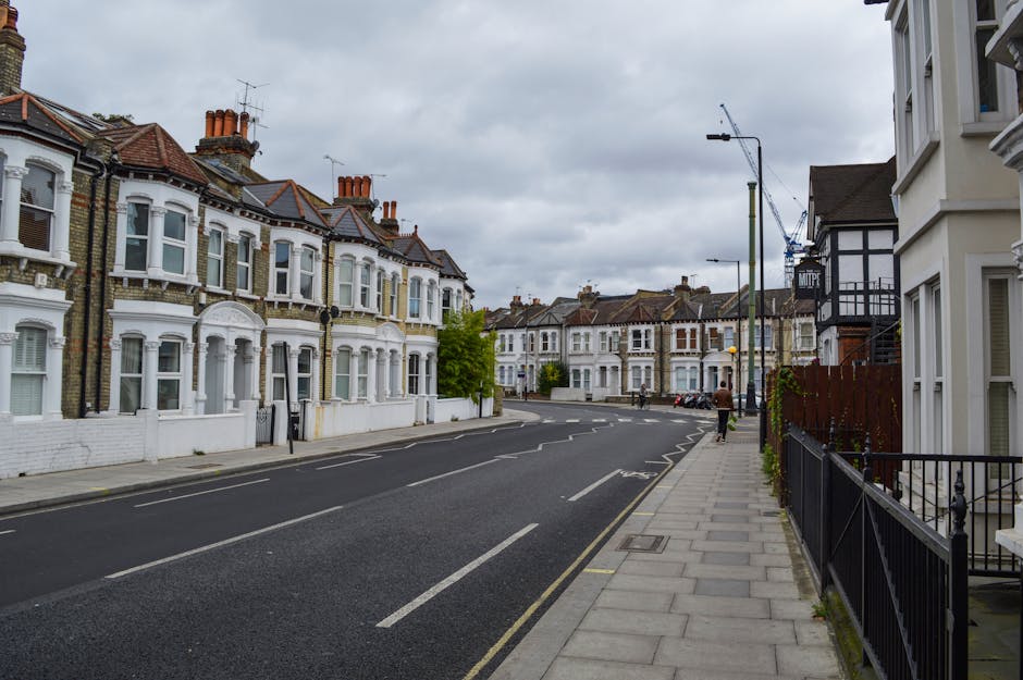A quiet residential street featuring a row of Victorian-style terraced houses with ornate white facades, decorative window frames, and brick chimneys, set against an overcast sky. The foreground shows a paved sidewalk lined with black metal railings and a few potted plants on the right side, while the street itself is marked with white dashed lines and parking bays, some occupied by parked cars. On the left, the houses have white-painted walls and red-tile roofs, with chimneys protruding above the rooftops. Tall streetlights and a crane are visible further down the road, indicating ongoing construction or maintenance. The scene captures a typical urban setting suitable for private rubbish clearance services by companies like Rubbish Clearance Fulham, highlighting the environment where on-site rubbish removal or alternative waste handling might be necessary. Overall, the image depicts a well-maintained, orderly neighborhood street during daytime with a neutral, professional tone, emphasizing its suitability for waste management operations within an urban residential context.