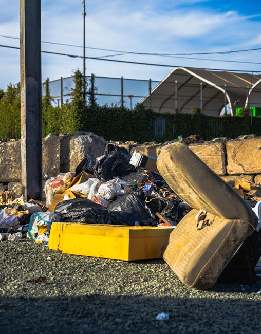 A close-up view of a roadside pile of mixed waste and rubbish, including black plastic garbage bags, discarded cardboard boxes, and a worn-out, beige car tyre leaning on some of the debris. The rubbish is spread on a rough asphalt surface with a yellow plastic container among the waste. In the background, there is a stone wall composed of irregularly shaped grey stones, partially obscuring a green hedge and trees behind it. Further out, a metal fence with netting attaches to the top of the wall, with a glimpse of a blue sky and some power lines overhead. The scene appears to depict an instance of private waste disposal, potentially handled by an independent rubbish removal service such as Rubbish Clearance Fulham, illustrating the type of unwanted waste that might require professional rubbish removal or on-site clearance services in Fulham and surrounding areas.
