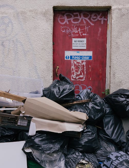 A collection of black garbage bags, cardboard, and debris piled against a rough-textured beige wall beneath a red metal door covered in graffiti and stickers, including one that reads 'Keep clear.' The bags and materials are placed on a paved surface, with some items visibly leaning or stacked, indicating an accumulation of waste awaiting collection. The door features various signs, including one stating 'No Money, No Exit,' and is surrounded by signs of neglect and graffiti, suggesting a site where independent rubbish clearance or non-standard waste disposal occurs. The scene is lit with natural daylight, highlighting the refuse's varying textures, from crumpled plastic to cardboard, and emphasizing the informal nature of waste accumulation in this urban environment, consistent with the type of rubbish removal services provided by Rubbish Clearance Fulham, especially in contexts involving private waste handling.