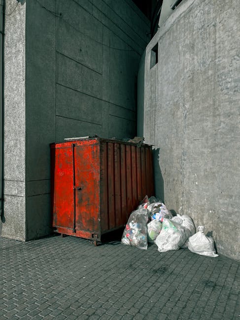 A large, rectangular metal skip in a deep red, weathered finish with visible rust and peeling paint, positioned against a dark green modern building façade. The skip is placed on a paved surface with a textured pattern of small, dark grey bricks. To its right, there are several transparent and white plastic rubbish bags filled with waste; some bags are tied closed, while others are partially open, revealing mixed household refuse inside. The skip is situated in a narrow alleyway or corner, bordered by a tall, smooth grey concrete wall on the right side and the building wall on the left, which features a small, square window or vent near the top. The scene appears to be outdoors, with an overcast ambient light creating a neutral, subdued atmosphere. The presence of the rubbish bags and skip indicates an ongoing or recent rubbish clearance, which aligns with private waste handling or onsite rubbish removal services, as provided by companies like Rubbish Clearance Fulham that specialise in alternative waste disposal solutions for property clearance or construction debris.