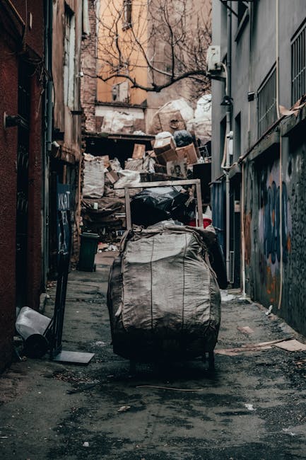 A collection of black garbage bags, cardboard, and debris piled against a rough-textured beige wall beneath a red metal door covered in graffiti and stickers, including one that reads 'Keep clear.' The bags and materials are placed on a paved surface, with some items visibly leaning or stacked, indicating an accumulation of waste awaiting collection. The door features various signs, including one stating 'No Money, No Exit,' and is surrounded by signs of neglect and graffiti, suggesting a site where independent rubbish clearance or non-standard waste disposal occurs. The scene is lit with natural daylight, highlighting the refuse's varying textures, from crumpled plastic to cardboard, and emphasizing the informal nature of waste accumulation in this urban environment, consistent with the type of rubbish removal services provided by Rubbish Clearance Fulham, especially in contexts involving private waste handling.